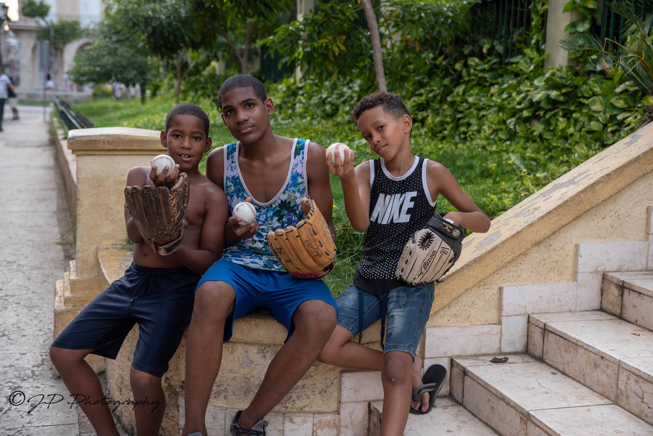 A picture of 3 children sitting on a wall, holding baseball equipment.